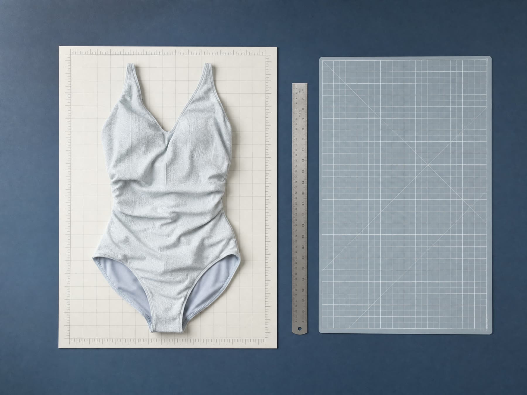 A swimsuit being laid out flat on a pale measuring mat beside a metal ruler, photographed from directly above.